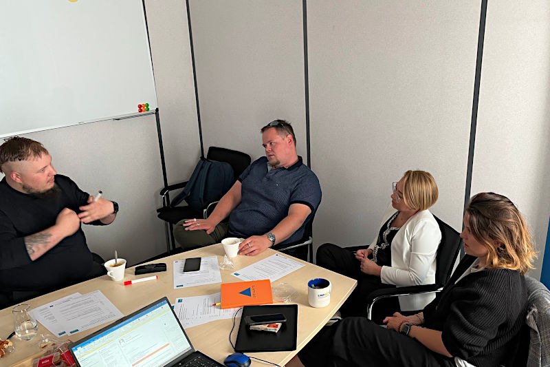 Two women and two men sit at a conference table and appear to be discussing something. There are notebooks and papers on the table and there's a whiteboard on the wall in the background.